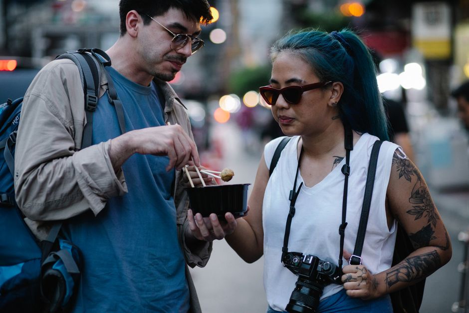 Two travelers savoring street food at night in a bustling urban setting.