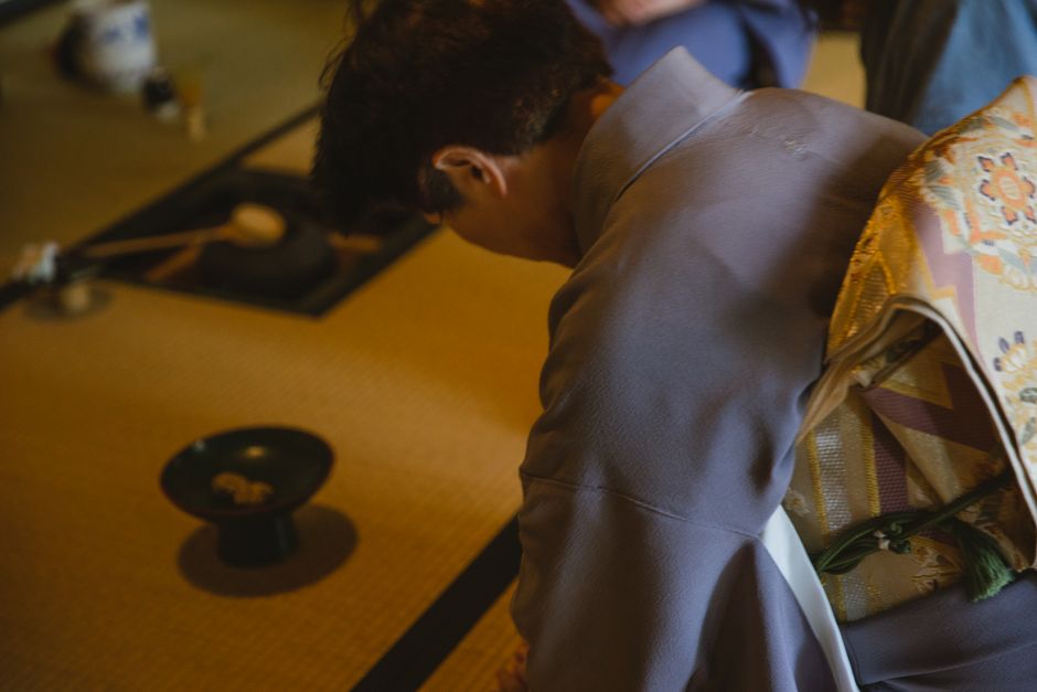 Side view of anonymous male in authentic robe bowing head while sitting on tatami with dishware for traditional tea ceremony